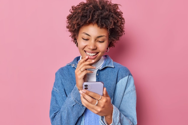 Captura interior de una guapa morena rizada mira la pantalla del teléfono inteligente y lee elogios debajo de su publicación vestida con una camisa de mezclilla se siente feliz aislada sobre fondo rosa concepto de tecnología