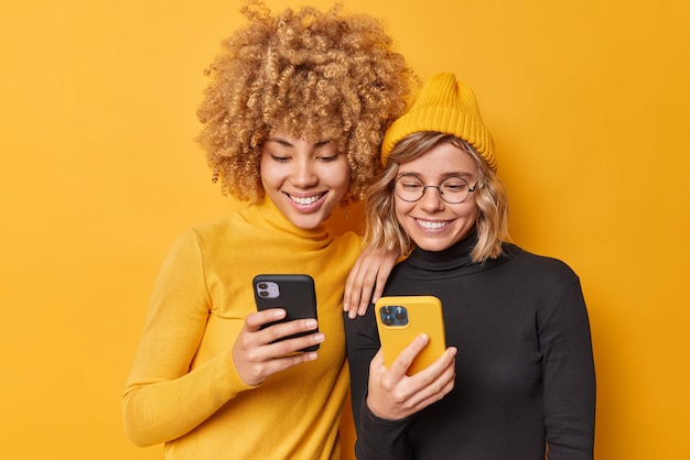 Captura interior de hermosas mujeres felices que disfrutan de una oferta móvil para navegar por una nueva aplicación o un sitio web que es adicto a las tecnologías modernas y mantiene el chat de celulares en línea aislado sobre un fondo amarillo vívido