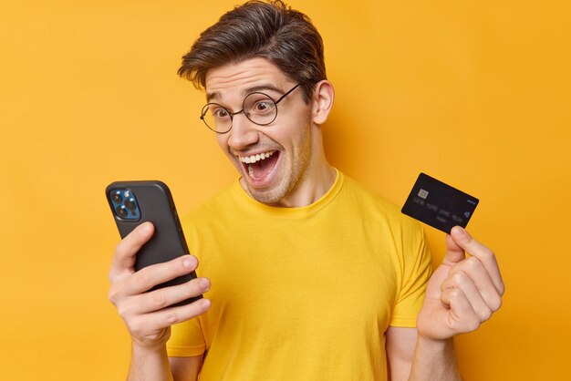 Captura interior de un hombre positivo que compra algo en línea sostiene una tarjeta de plástico del teléfono móvil feliz de obtener una suma global de dinero en su cuenta bancaria usa anteojos redondos y una camiseta casual aislada en la pared amarilla