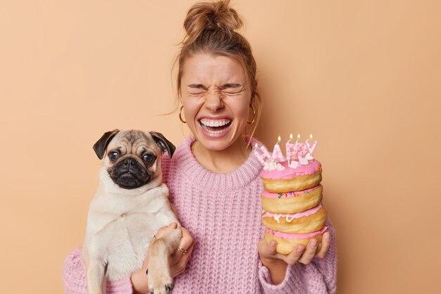 Captura interior de una joven emocional que exclama en voz alta que se prepara para la celebración del cumpleaños de las mascotas sostiene un perro pug pedigrí y donuts festivos con velas encendidas aisladas sobre fondo marrón