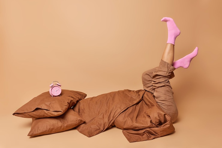 Foto gratuito captura interior de una mujer irreconocible vestida con pijama que mantiene las piernas levantadas usa calcetines posa en la cama debajo de una manta no quiere levantarse aislada sobre el fondo beige del estudio duro despertar