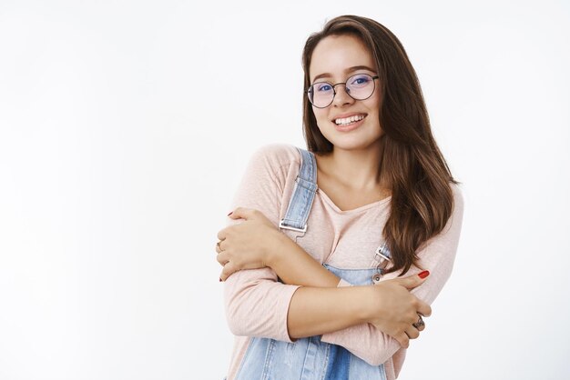 Captura de pantalla de una linda y amable mujer de aspecto amigable de 20 años con anteojos transparentes abrazándose con las manos cruzadas sobre el cuerpo sintiéndose fría y sonriendo a la cámara sobre un fondo gris