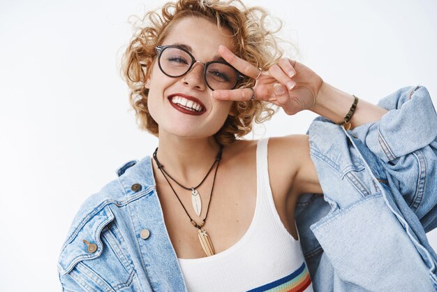 Captura de pantalla de una mujer carismática y alegre despreocupada que disfruta de la vida mostrando el signo de la paz o la victoria sobre la cabeza inclinada del ojo emocionada y complacida sonriendo ampliamente divirtiéndose sobre fondo blanco