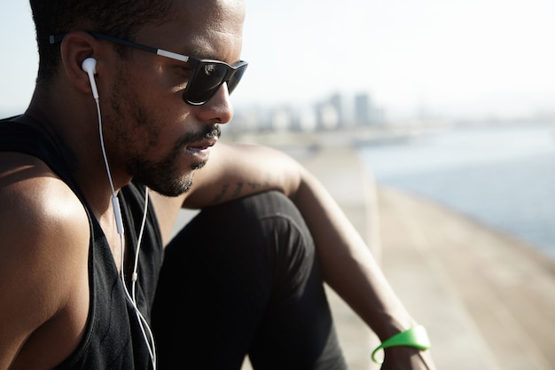 Captura recortada de pensativo joven apuesto basculador con gafas de sol sentado en la cima de las escaleras de piedra, vestido con ropa deportiva negra, triste y serio, escuchando música en su teléfono inteligente, descansando