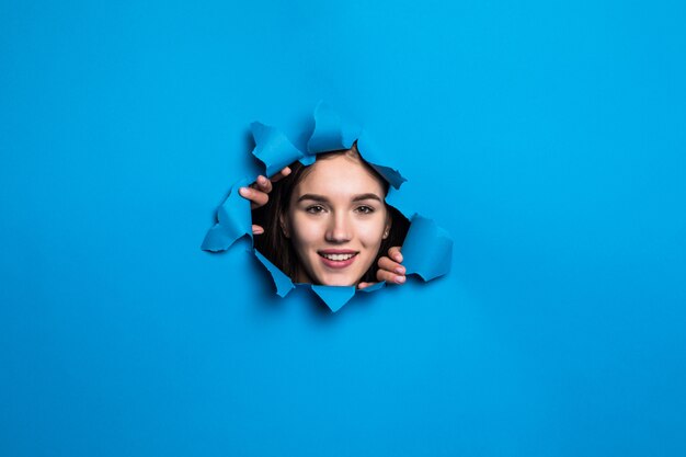 Cara bonita joven mirando a través del agujero azul en la pared de papel.