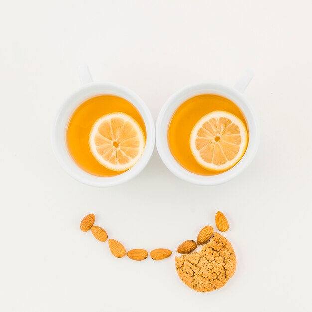 Cara feliz hecha con taza de té de limón; Almendras y galletas comidas sobre fondo blanco