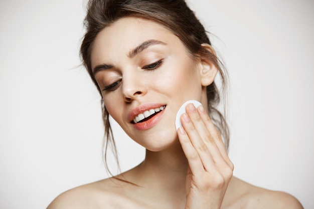 Cara de limpieza hermosa chica morena natural con esponja de algodón sonriendo sobre fondo blanco. Cosmetología y spa.