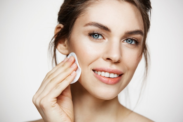 Cara de limpieza hermosa chica morena natural hermosa con esponja de algodón sonriendo mirando a cámara sobre fondo blanco. Cosmetología y spa.