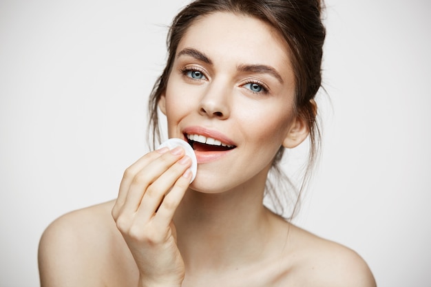 Cara de limpieza hermosa chica morena natural hermosa con esponja de algodón sonriendo mirando a cámara sobre fondo blanco. Cosmetología y spa.