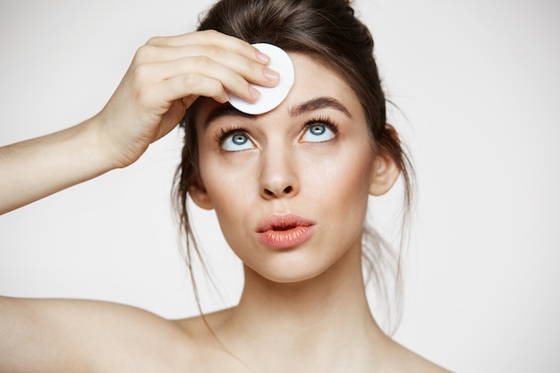 Cara de limpieza hermosa chica morena natural hermosa con esponja de algodón sonriendo sobre fondo blanco. Cosmetología y spa.