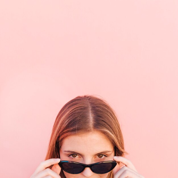 La cara de la mujer joven que mira a escondidas a través de gafas de sol negras contra el contexto rosado