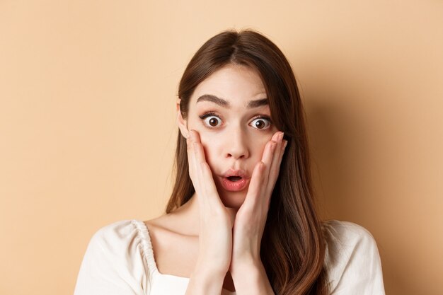 Cara de niña sorprendida diciendo wow tocando las mejillas y mirando emocionado a la cámara de pie sobre un fondo beige ...