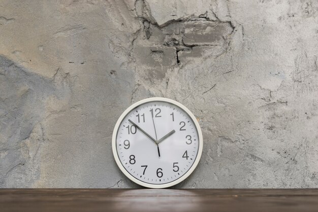 Cara de reloj redonda que se inclina contra el muro de cemento dañado en el escritorio de madera