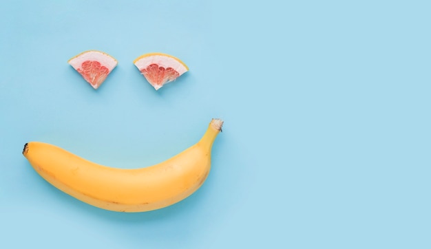 Cara sonriente hecha con plátano amarillo y rodaja de pomelo sobre fondo azul