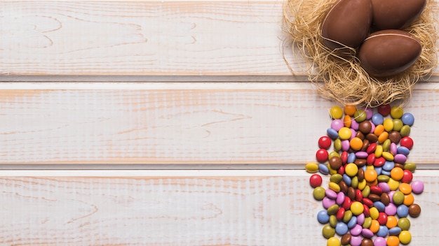 Foto gratuita caramelos de gemas multicolores cerca de los huevos de chocolate en el nido sobre el escritorio de madera