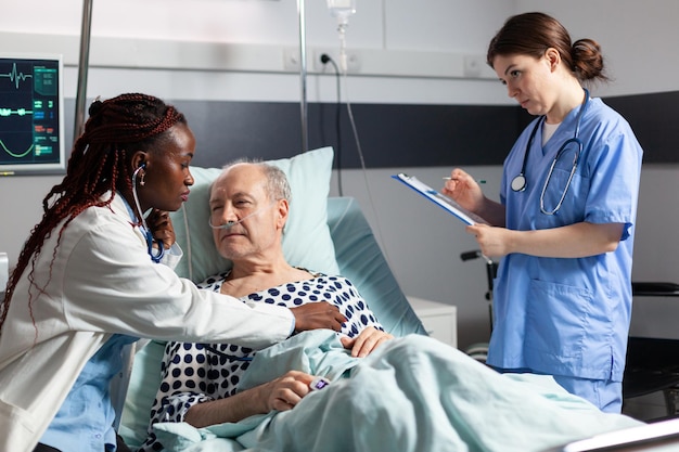 Cardiólogo afroamericano revisando el corazón del paciente mayor, usando estetoscopio mientras el paciente está acostado en la cama del hospital para establecer el diagnóstico para la terapia, respirando con la ayuda de un tubo de ensayo.