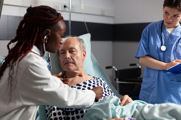 Cardiólogo joven que revisa el corazón del paciente mayor que examina, usando el estetoscopio mientras el paciente está acostado en la cama del hospital para establecer el diagnóstico para la terapia, respirando con la ayuda del tubo de ensayo