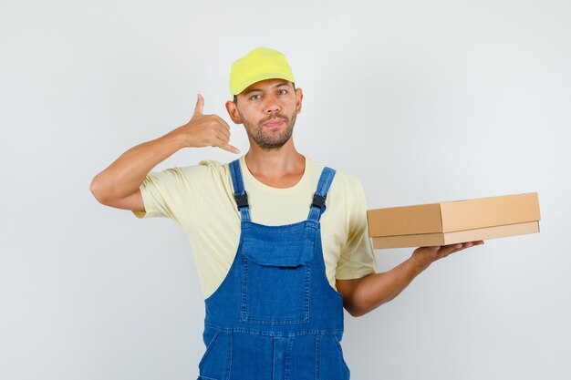 Cargador joven con caja de cartón con gesto de teléfono en uniforme, vista frontal.