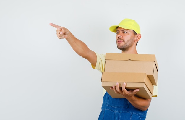 Cargador joven sosteniendo cajas de cartón y apuntando hacia afuera en uniforme, vista frontal.