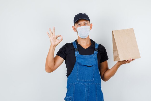 Cargador joven en uniforme, máscara con bolsa de papel con signo de ok, vista frontal.