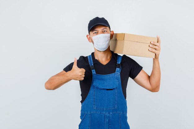 Cargador joven en uniforme, máscara con caja de cartón con el pulgar hacia arriba, vista frontal.