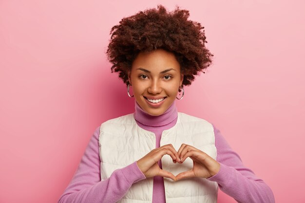 Cariñosa dama afroamericana muestra gesto de corazón, expresa amor, admiración y simpatía, sonríe feliz, muestra dientes blancos, demuestra afecto, usa chaleco blanco.