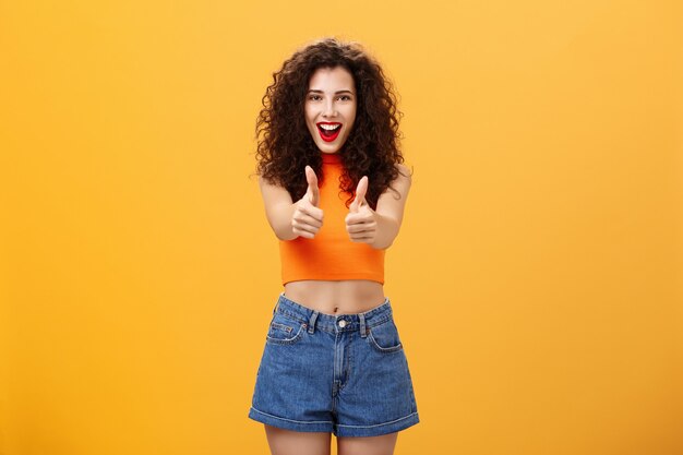 Carismática, ambiciosa y extrovertida mujer caucásica encantadora con peinado rizado y lápiz labial rojo que muestra los pulgares para arriba gesto de aprobación o aprobación sonriendo alegremente apoyando sobre la pared naranja.