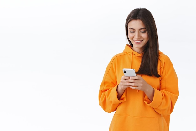 Carismática chica morena bonita en sudadera con capucha naranja sonriendo y mirando la pantalla del teléfono móvil como mensajes de texto, mensajes con su novio, usando la aplicación de teléfono