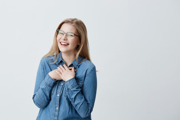 Carismática y encantadora joven europea con cabello rubio lacio, con elegantes gafas y camisa vaquera, sonriendo ampliamente, esperando sorpresa, luciendo feliz