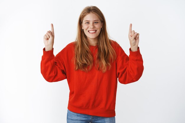 Carismática, encantadora y optimista joven europea con mejillas regordetas, ojos azules y pecas sonriendo alegremente levantando las manos para señalar el espacio de la copia posando en un elegante suéter rojo sobre una pared gris