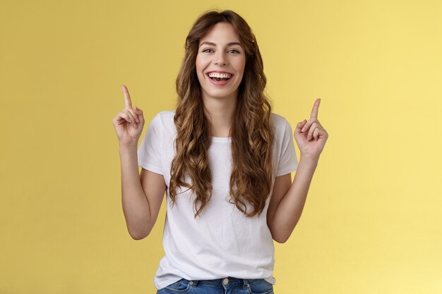 Carismática, entusiasta, atractiva mujer animada que se divierte disfrutar de una increíble noche de fiesta riendo alegremente apuntando hacia arriba con los dedos índices para introducir promo sonriendo felizmente fondo amarillo