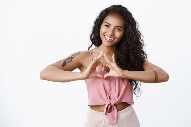 Foto gratuita carismática mujer linda con peinado rizado, inclinar la cabeza y reír con una sonrisa blanca con dientes, mostrar el signo del corazón expresar amor, conmovedores sentimientos conmovedores, pared de estudio