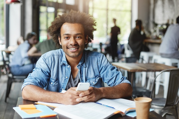 Carismático y guapo estudiante universitario afroamericano con barba mediante conexión inalámbrica a Internet en su dispositivo electrónico durante el almuerzo