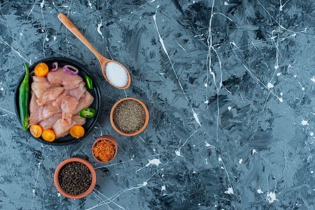 Carnes y verduras marinadas en un plato junto a las especias en tazones y cuchara, sobre el fondo azul.