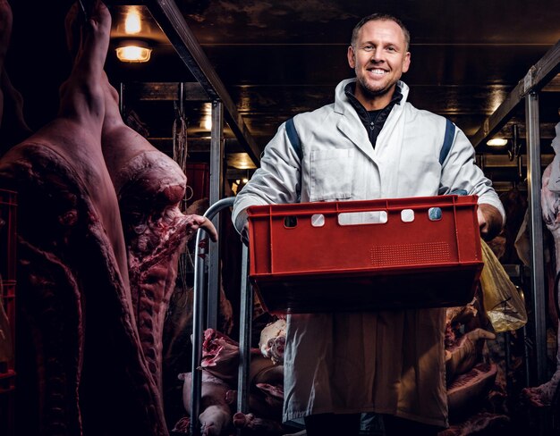 Carnicero alegre en ropa de trabajo sosteniendo una caja con trozos de carne mientras está de pie en medio de canales de carne