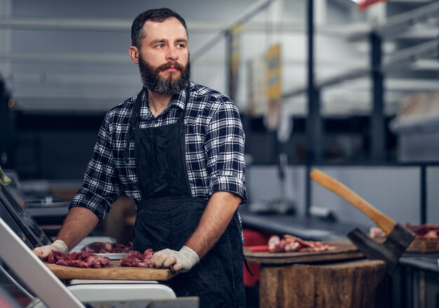 Carnicero barbudo vestido con una camisa polar que sirve carne fresca cortada en un mercado.