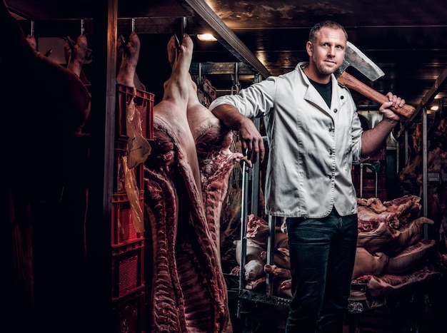 El carnicero con una camisa de trabajo blanca sosteniendo un hacha mientras está parado en un almacén refrigerado en medio de canales de carne