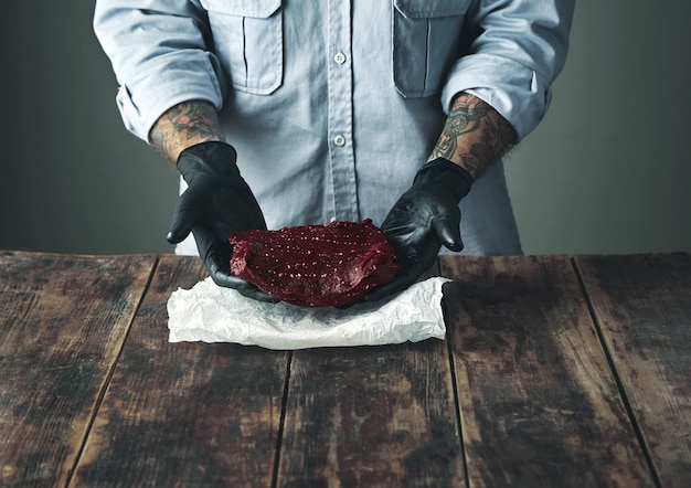Carnicero tatuado irreconocible con guantes negros ofrece un trozo de carne de ballena de lujo sobre papel artesanal blanco