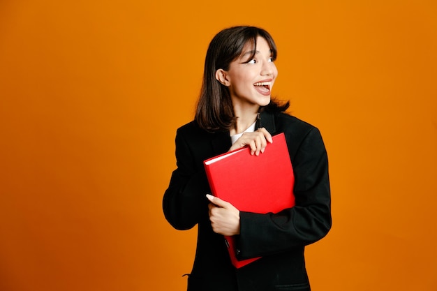 Carpeta de tenencia sonriente con pluma joven hermosa mujer vistiendo chaqueta negra aislado sobre fondo naranja