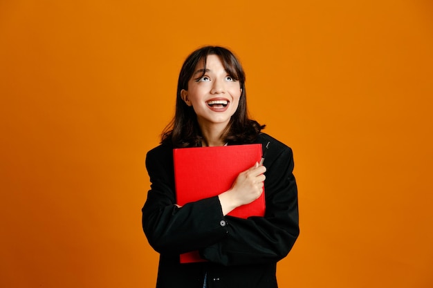 Carpeta de tenencia sonriente con pluma joven hermosa mujer vistiendo chaqueta negra aislado sobre fondo naranja