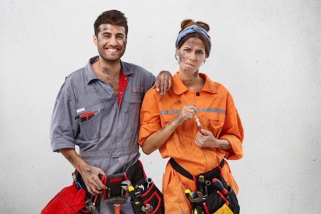 Carpintero femenino cansado en uniforme naranja, tiene destornillador y colega masculino con expresión feliz