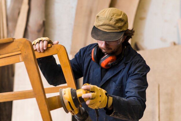 Carpintero profesional que pule una silla de madera con una lijadora eléctrica.