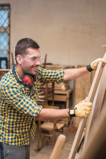 Foto gratuita carpintero de sexo masculino sonriente que lleva los guantes protectores que sostienen el tablón de madera en el taller