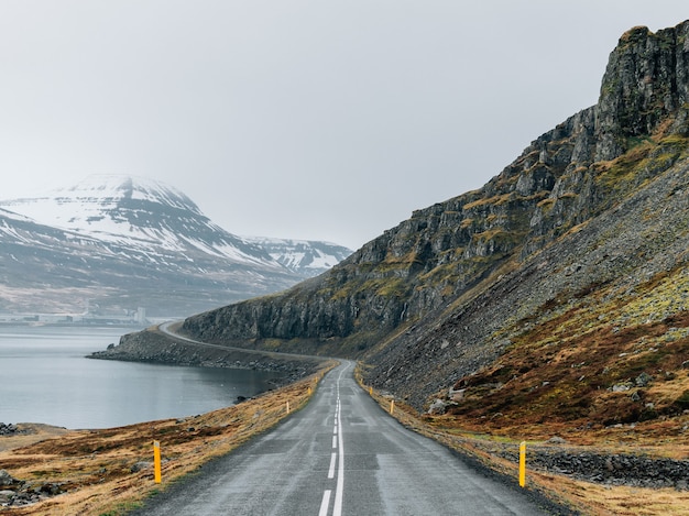 Carretera con curvas rodeada por el mar y rocas cubiertas de vegetación y nieve bajo un cielo nublado