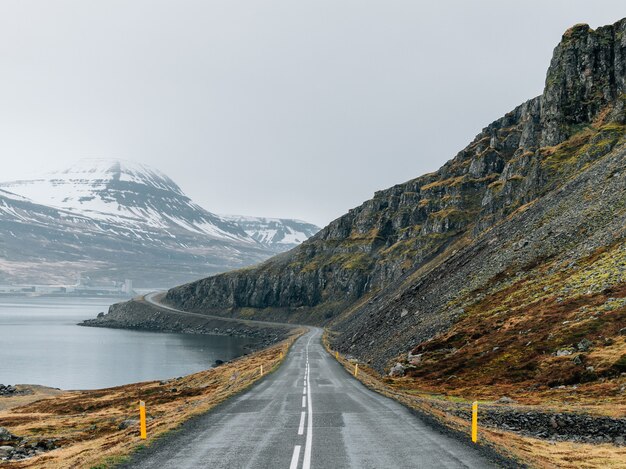 Carretera con curvas rodeada por el mar y rocas cubiertas de vegetación y nieve bajo un cielo nublado