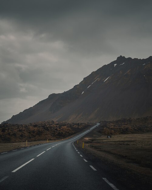 Carretera vacía con curvas junto a una hermosa montaña rocosa bajo un cielo gris y sombrío