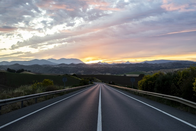 Carretera vacía rodeada de colinas bajo el cielo nublado del atardecer