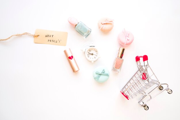 Carrito de compras con reloj despertador pequeño, macarrones, etiqueta de venta, lápiz labial y esmalte de uñas
