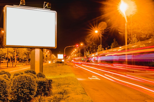 Cartelera en blanco blanco cerca de la carretera con senderos de luz de tránsito de hora punta