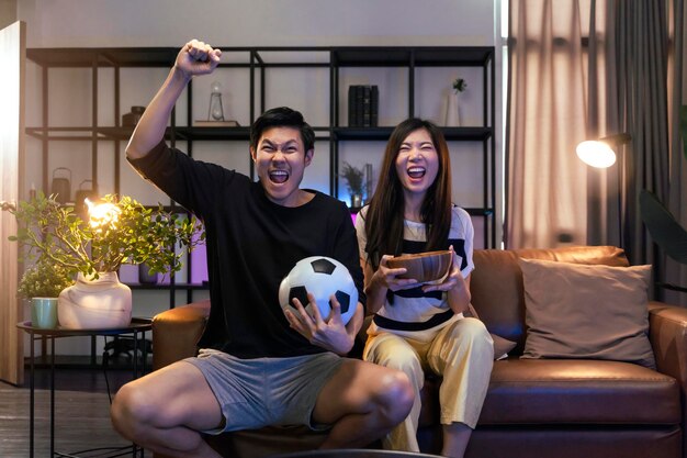Casarse con adultos asiáticos viendo la televisión en casa Pareja asiática animando la competencia de juegos deportivos junto con risa sonrisa victoria en el sofá sofá en la sala de estar actividad de aislamiento en el hogar
